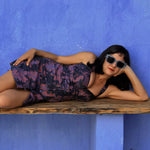 Load image into Gallery viewer, A woman wearing a dark floral halter dress and sunglasses reclines on a rustic wooden ledge against a textured blue wall.
